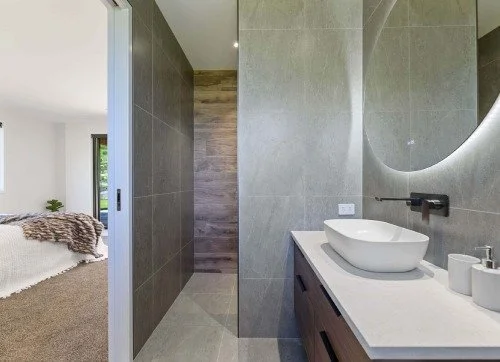 Modern bathroom with gray tiled walls, a wooden vanity with a vessel sink, circular mirror, and minimal accessories.