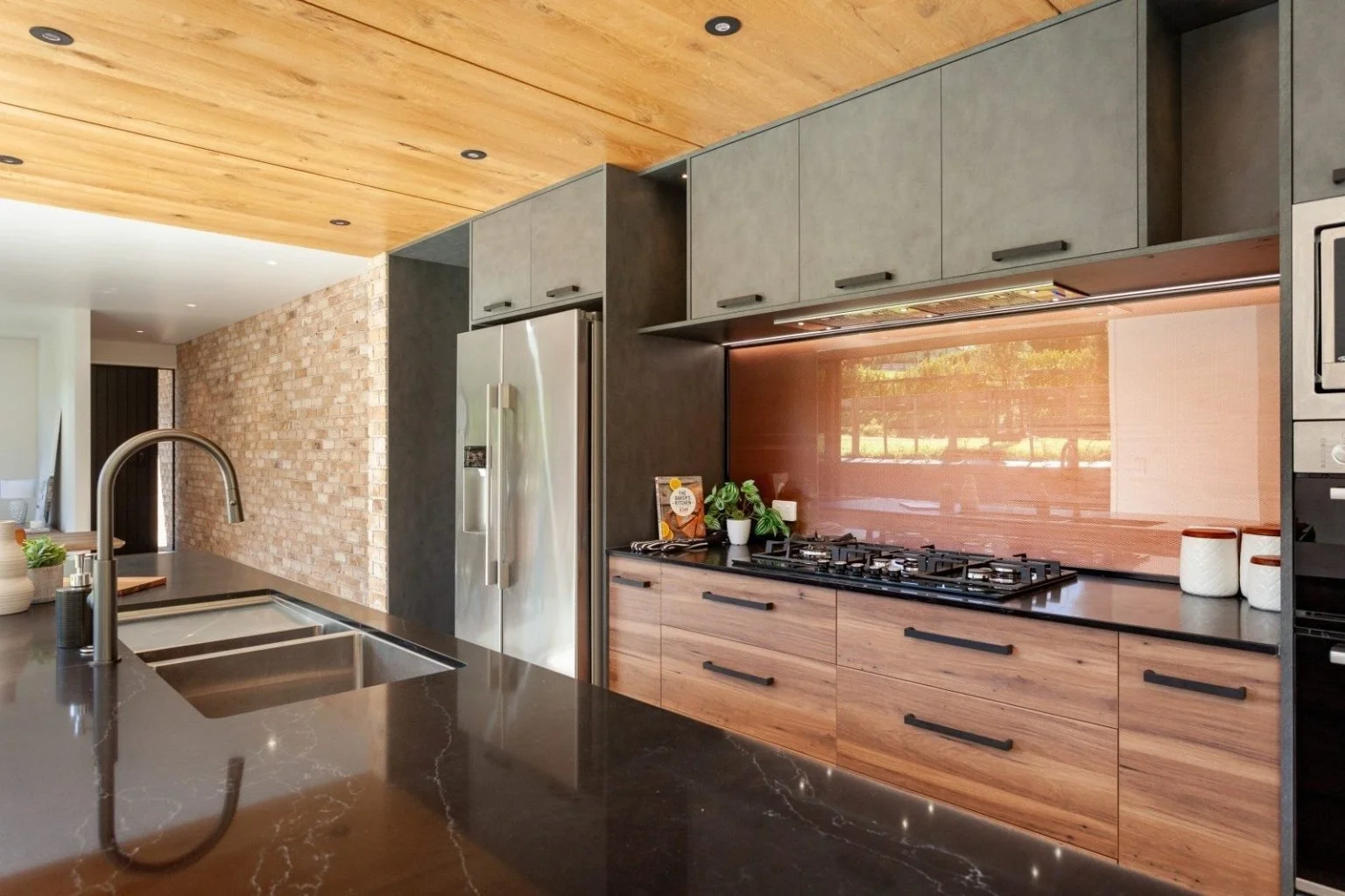 Modern kitchen with dark countertops, wooden cabinetry, stainless steel appliances, a brick wall, and a wooden ceiling.