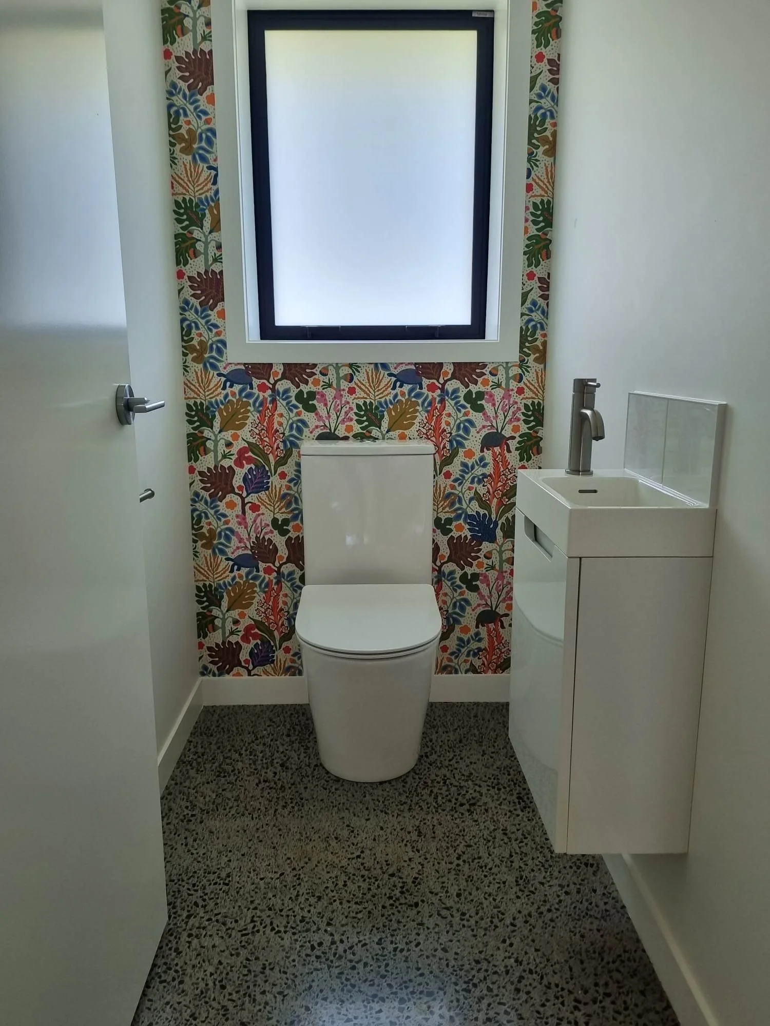 Small bathroom with a patterned wallpaper behind the toilet, a window above the toilet, and a white sink with a modern faucet on the right. The floor has a speckled tile pattern.