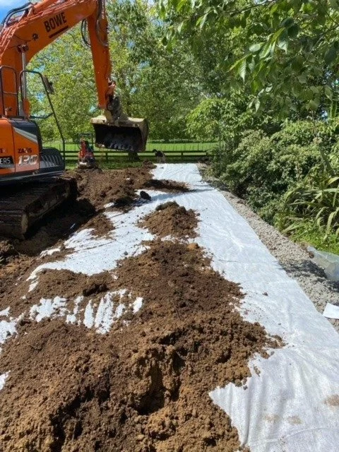 An orange excavator is working on a dirt path lined with white fabric, with piles of dirt along the path's edge. Green trees and bushes are visible in the background.
