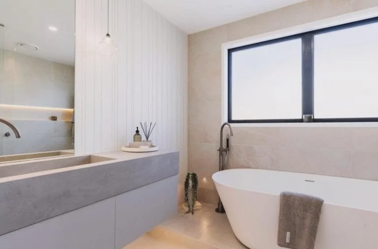 Modern bathroom featuring a large window, a white bathtub with a gray towel, a cactus plant, and a countertop with toiletries.