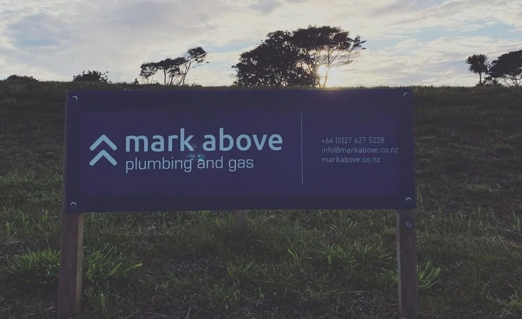 A purple sign for 'mark above plumbing and gas' on a grassy area at sunset, with trees and the sun in the background.