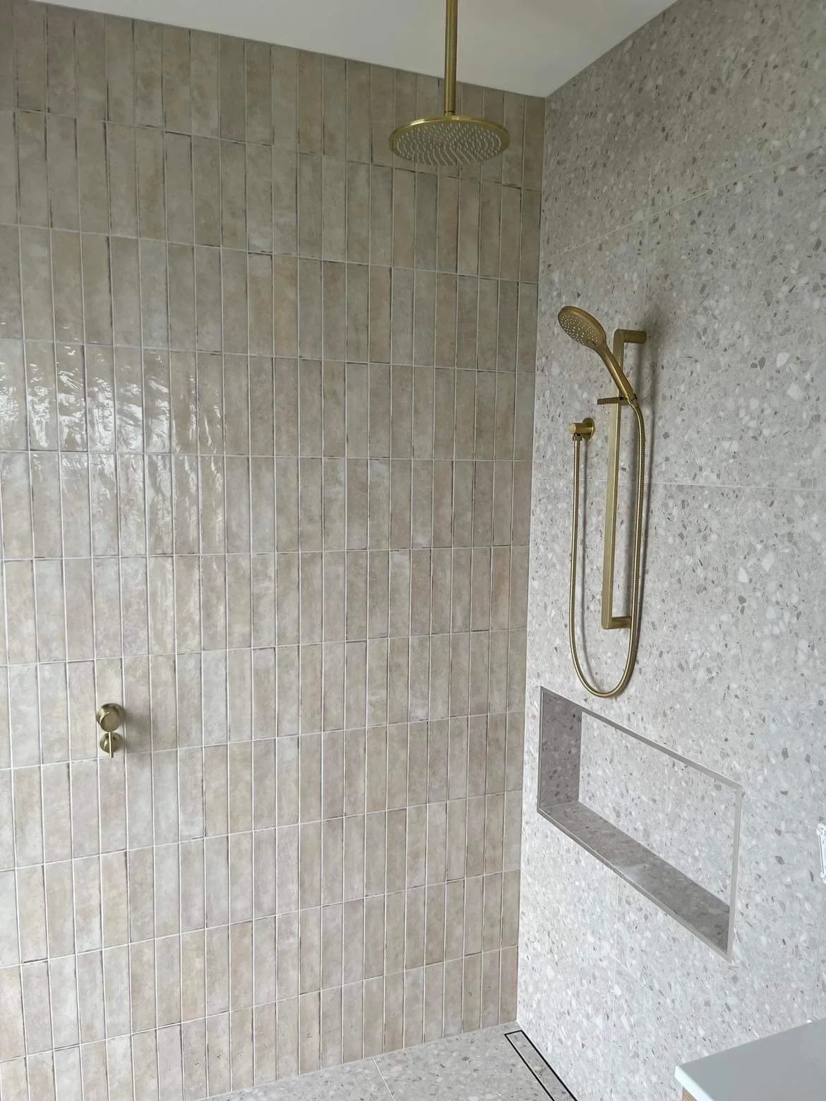 Modern shower with beige tiled walls, a gold overhead rain shower, a handheld showerhead, and a built-in niche for toiletries.