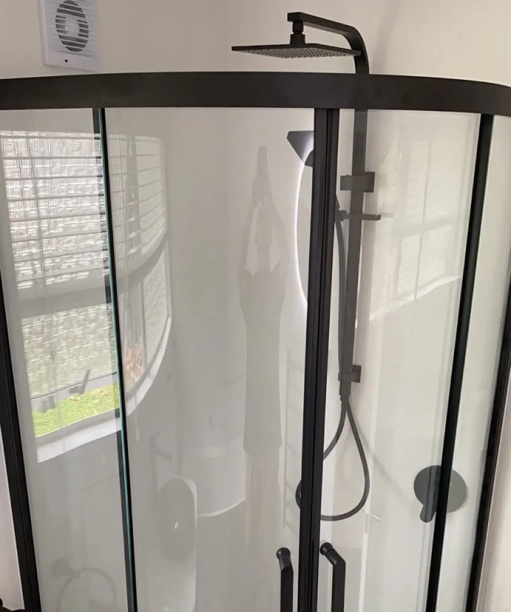 Modern shower enclosure with glass doors, black frame, overhead rain shower, handheld shower wand, and wall-mounted shower bar.