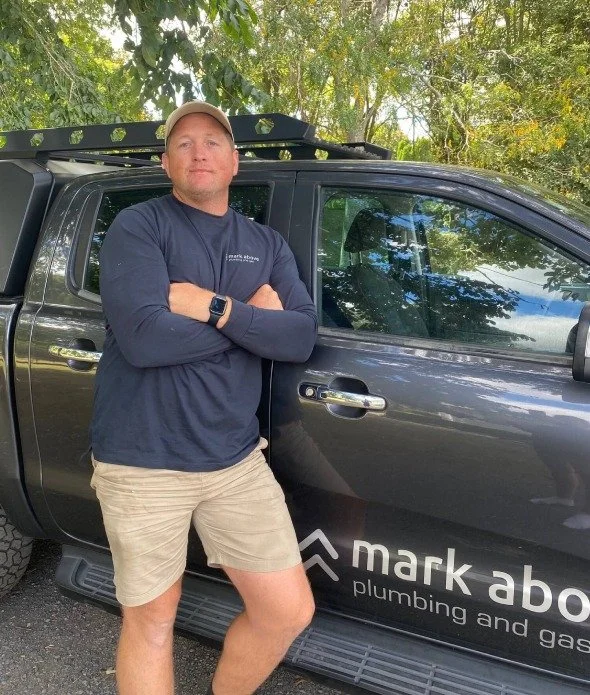 A man standing with arms crossed next to a black pickup truck, outdoors with trees in the background.