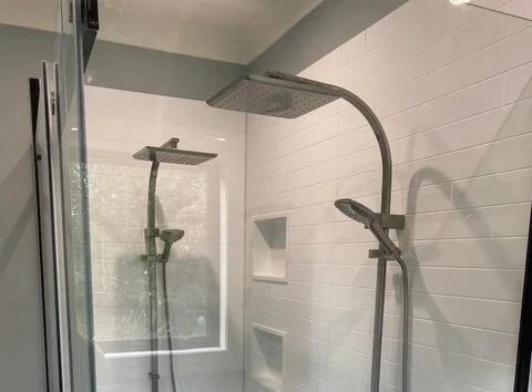 Two shower heads with handheld attachments in a modern shower with white tiled walls.