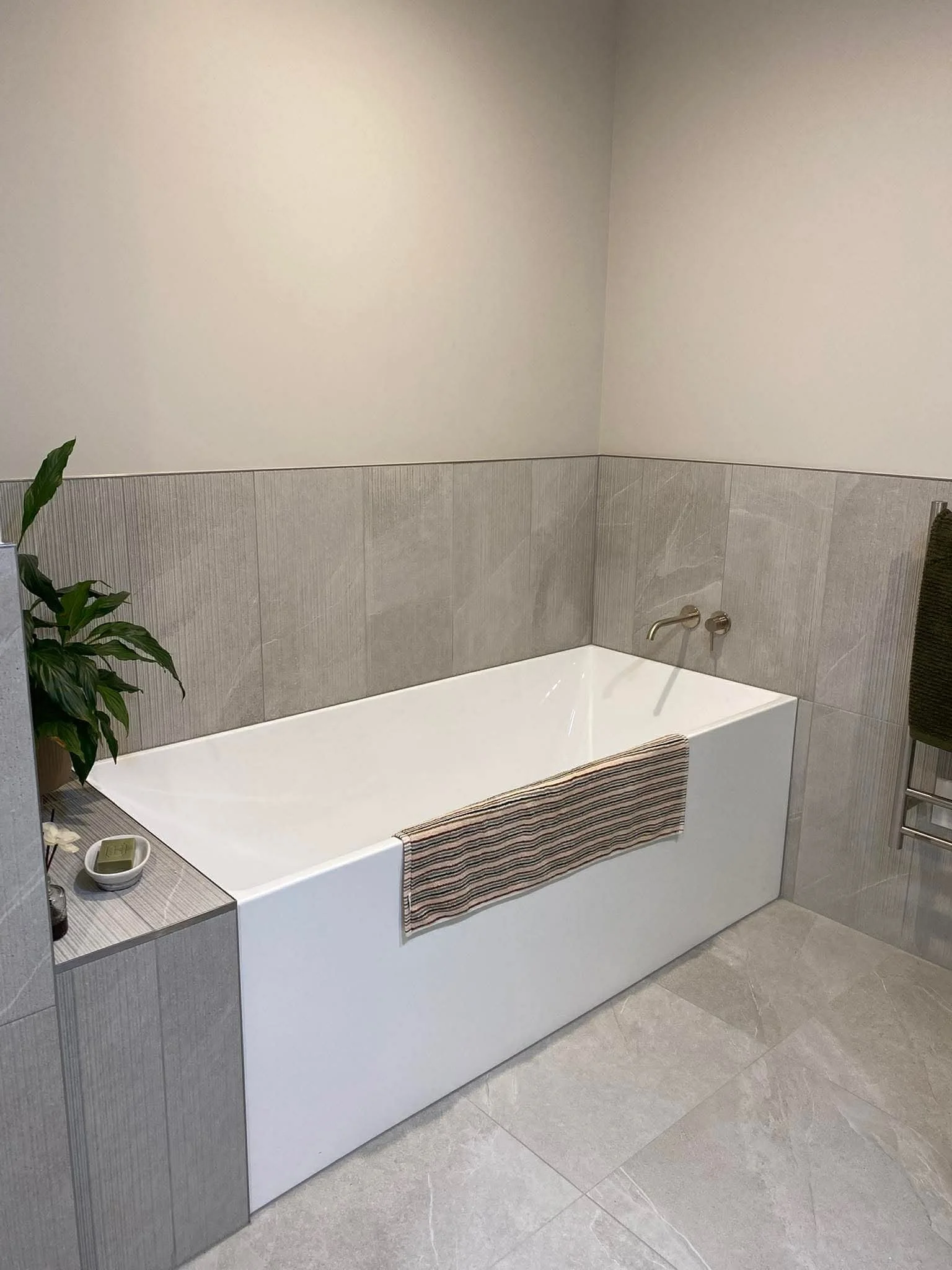 Bathroom with a white bathtub, a striped bath towel hanging on the side, a potted plant on a side table, and a beige tiled wall.