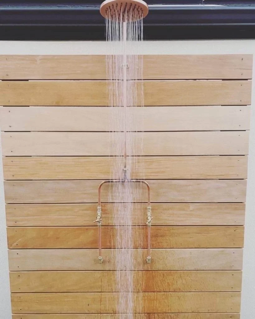 A wooden wall with a copper pipe and a round showerhead, with water cascading from the showerhead.