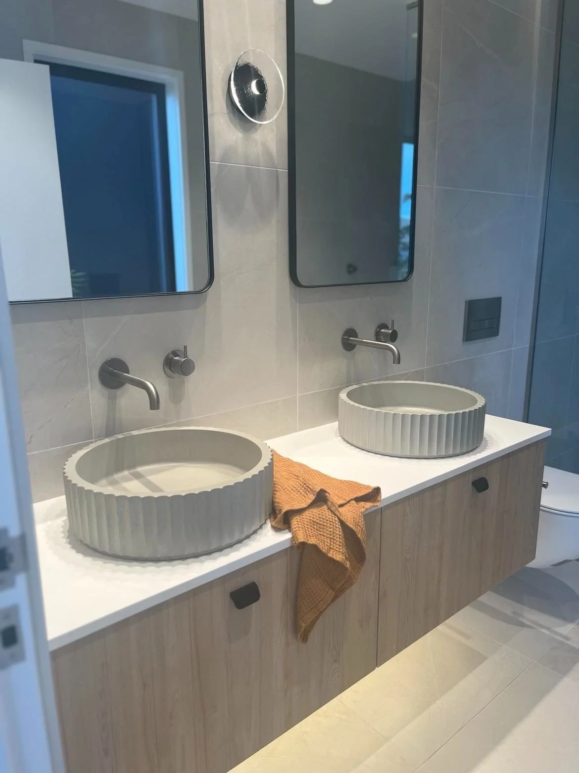 Modern bathroom with two round vessel sinks on a white countertop, brown towel between them, two mirrors above, and gray wall tiles.
