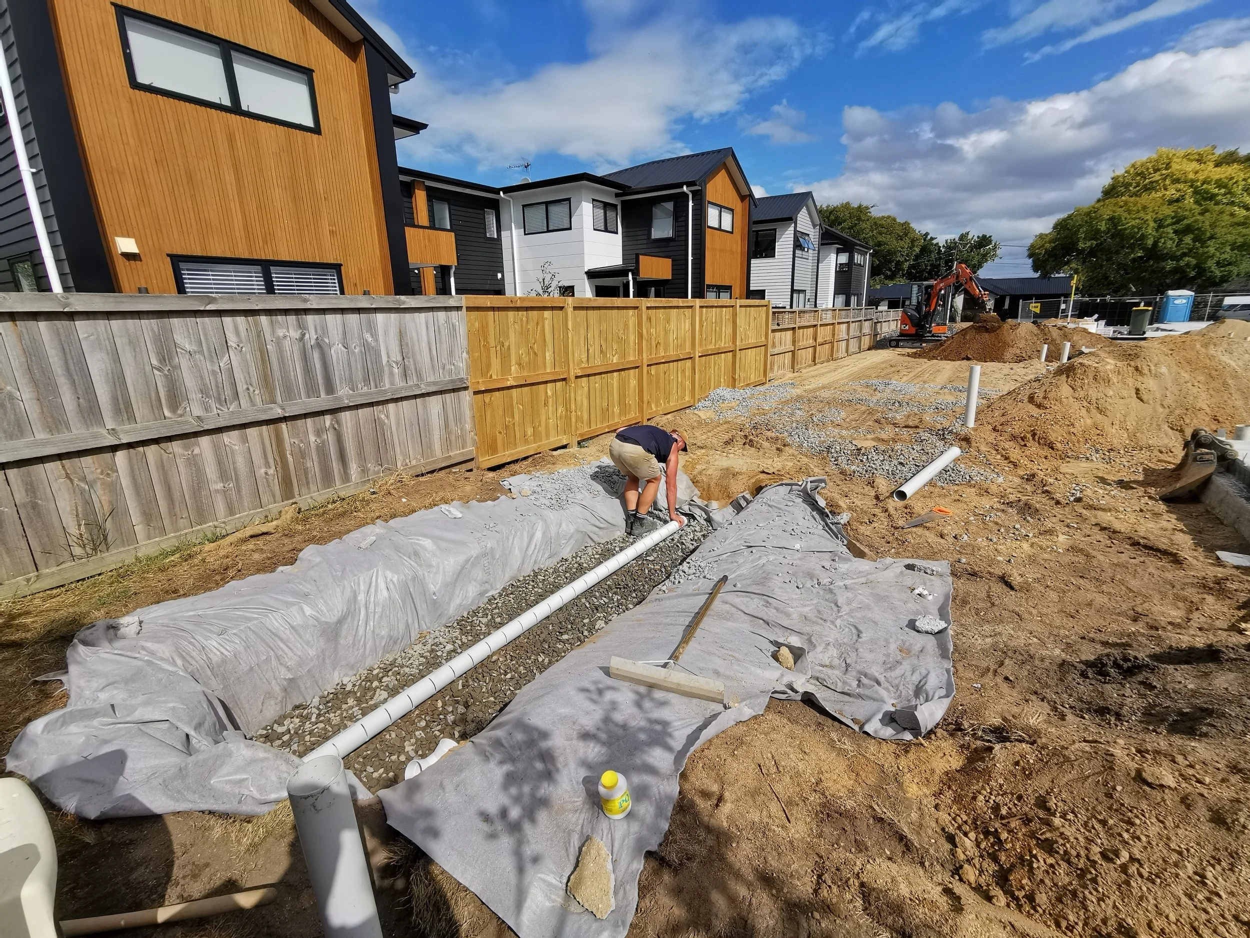 Construction site in a residential area with new homes and a worker installing PVC pipes in the ground, surrounded by dirt, gravel, and construction materials.