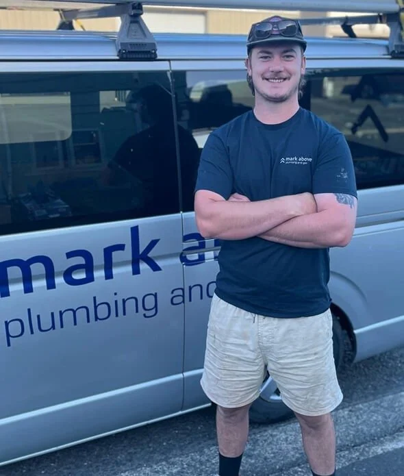 A smiling young man with crossed arms standing in front of a company van with the words "mark above plumbing and" visible on the side. He is wearing a navy T-shirt, beige shorts, and sunglasses on his head.