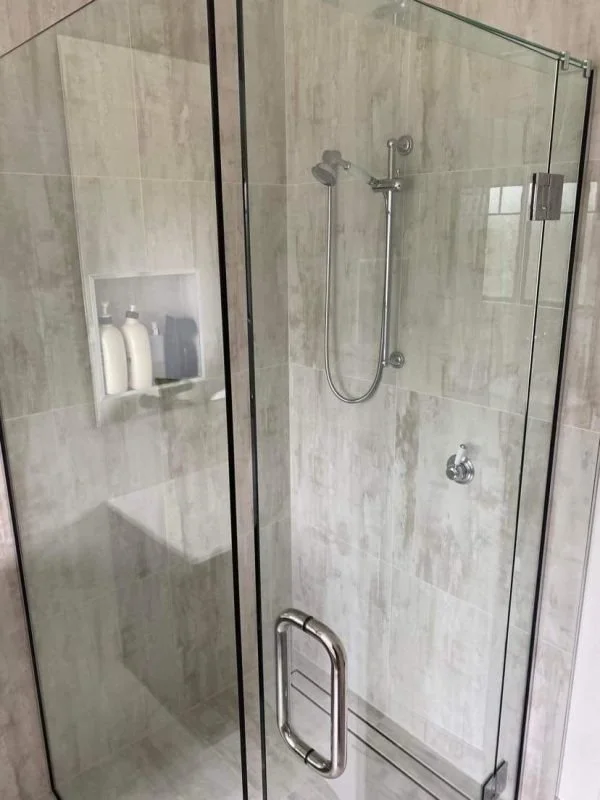 Inside a glass-enclosed shower with beige tiles, a mounted showerhead, and a built-in niche with bottles.