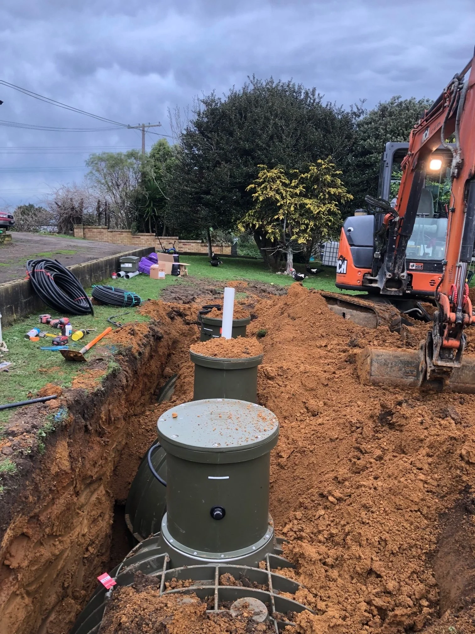 Excavation site with three underground utility vaults installed in a deep trench, construction tools, pipes, and an orange excavator on cloudy day.