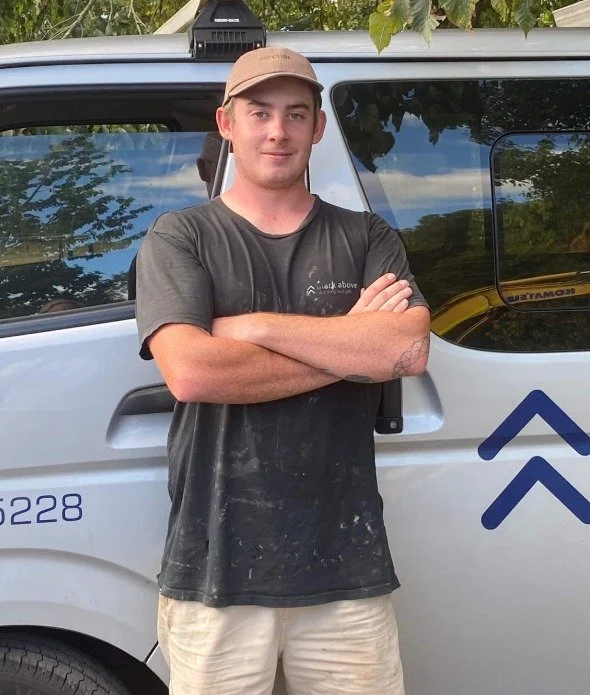 A young man with crossed arms standing in front of a silver vehicle with company logos, wearing a beige cap and a dark shirt, outdoors with trees reflected in the vehicle's windows.