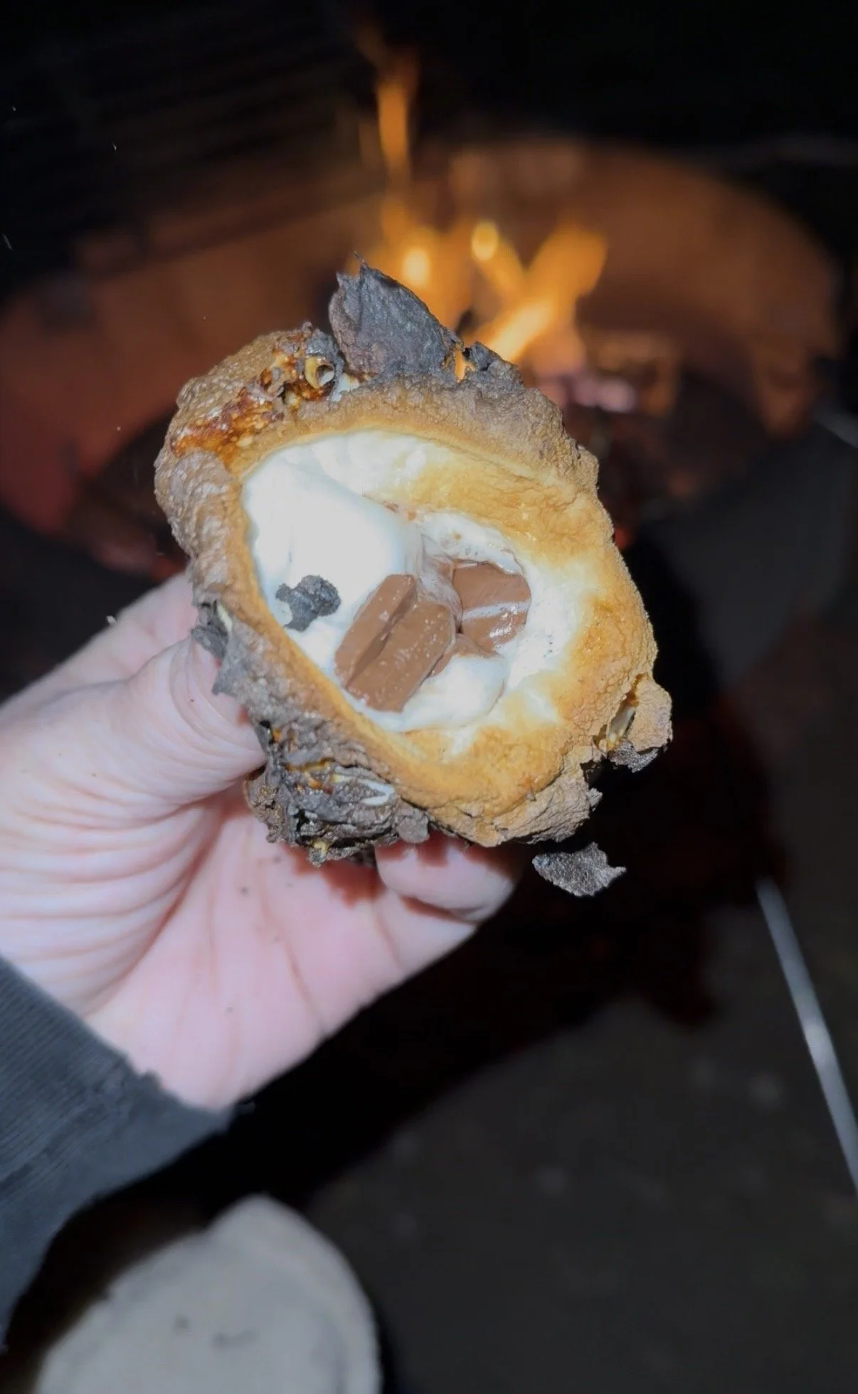 Person holding a roasted chili pepper filled with cheese and chocolate pieces, with a campfire in the background.