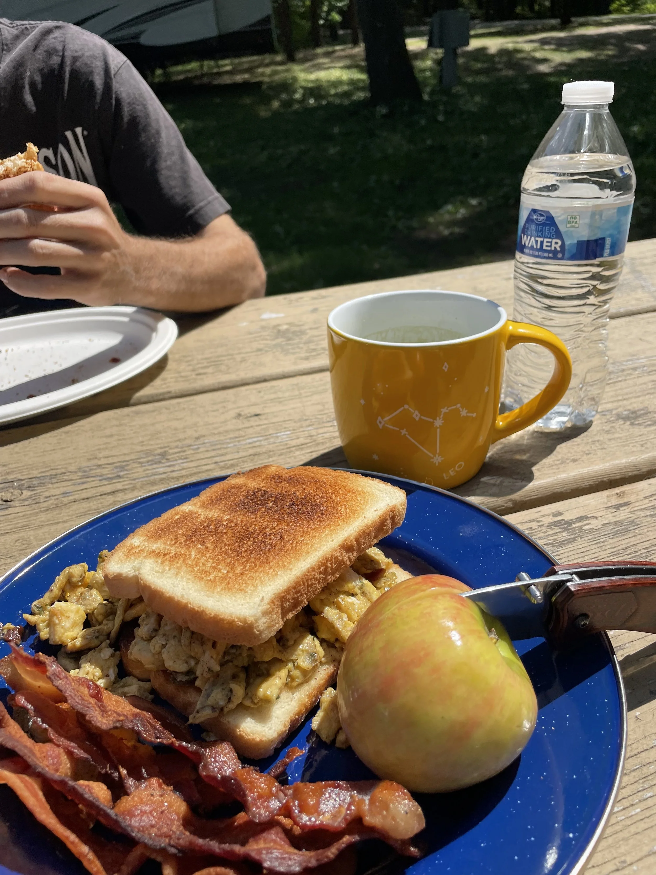 Breakfast on a wooden table includes a blue plate with a sandwich, bacon, and a green apple, a yellow mug with a constellation design, a bottle of water, and a person in the background holding a piece of toast.