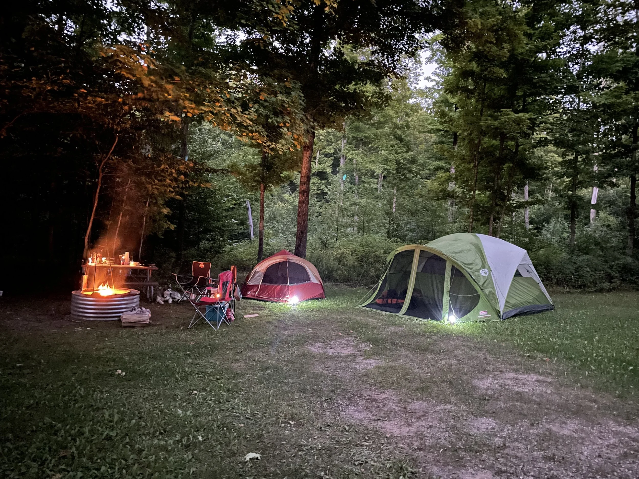 Campground scene at dusk with two tents, folding chairs, a small table, and a campfire in a wooded area.