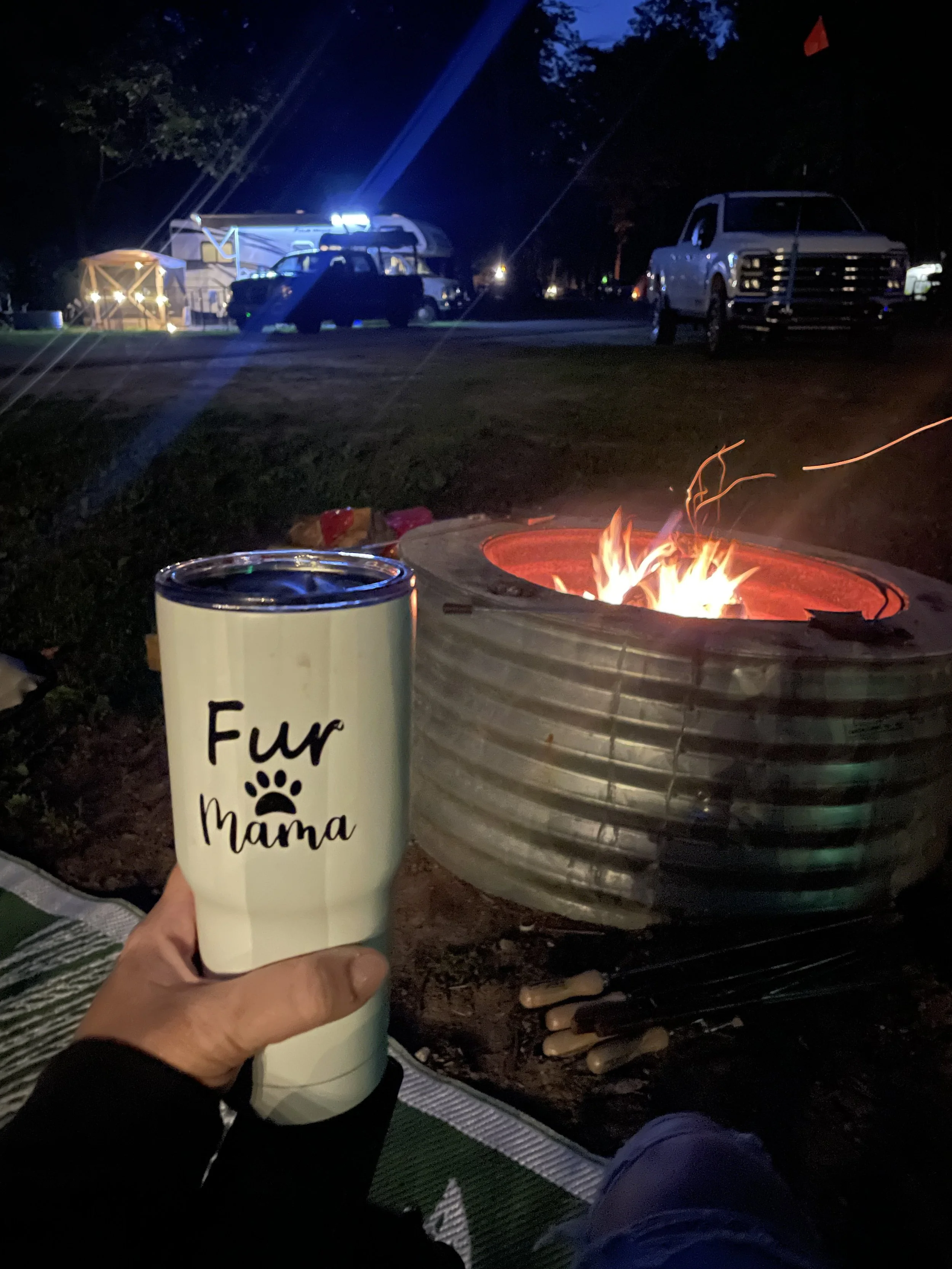 A person's hand holding a tumbler with the words "Fur Mama" written on it, sitting near a campfire at night. In the background, there are parked trucks and vehicles, and a camping setup with a tent and lights.