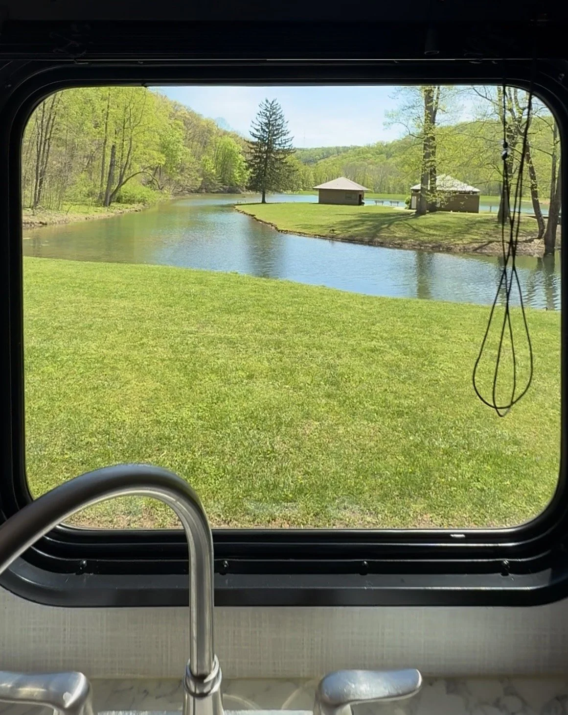 View through a window of a lake with green grass and trees, and small buildings in the background, with a faucet visible at the bottom of the window.