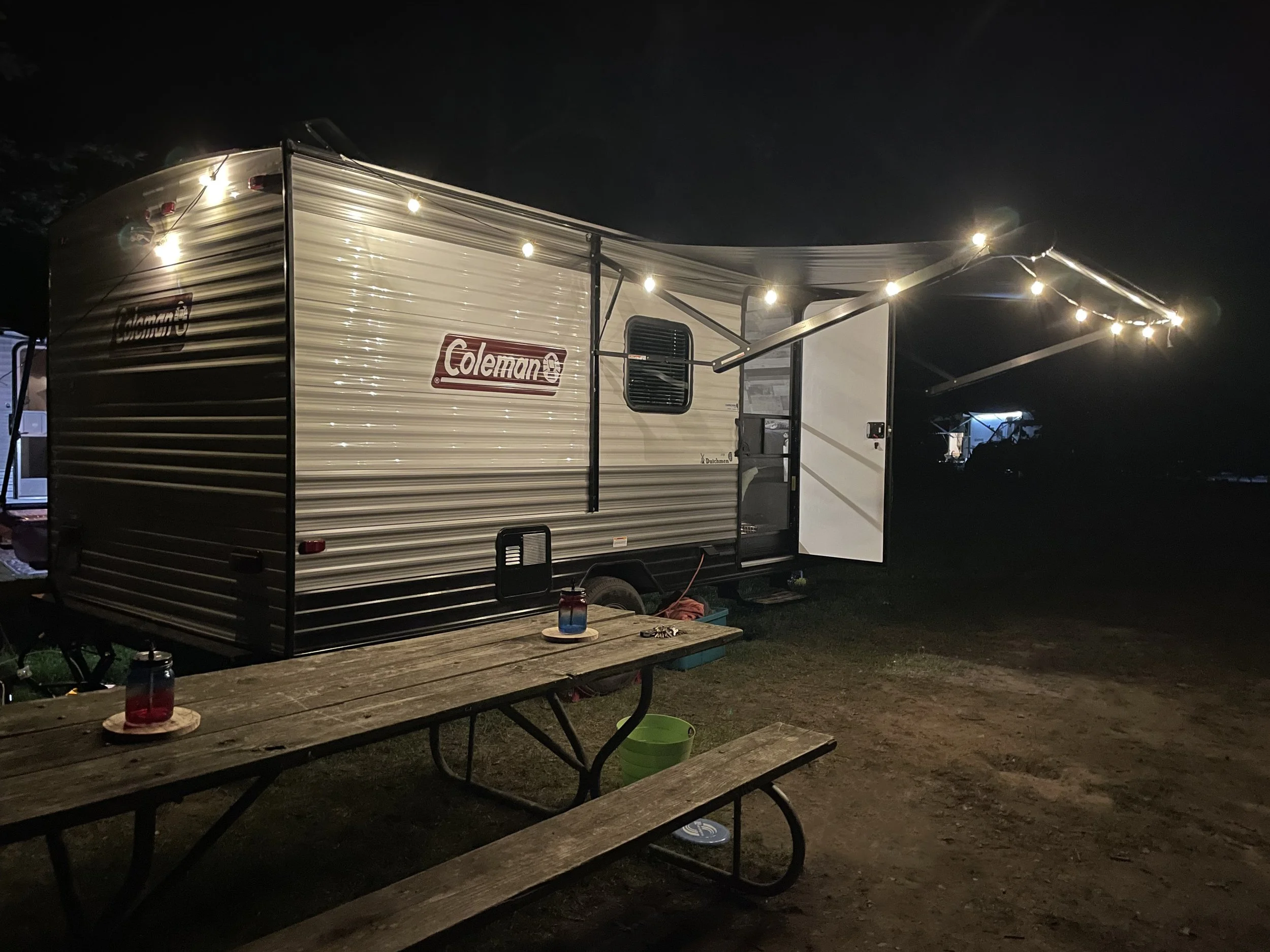 A Coleman travel trailer at night with string lights overhead, a picnic table outside, and a dark background.