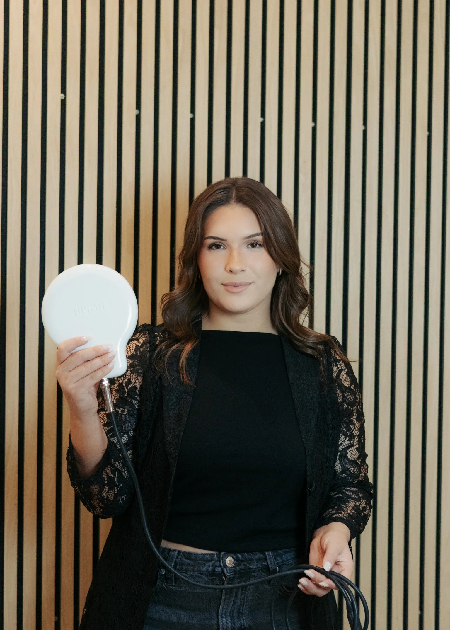 A young woman with brown hair wearing a black lace jacket and black top, holding a white electric device against a wooden vertical slat wall background.