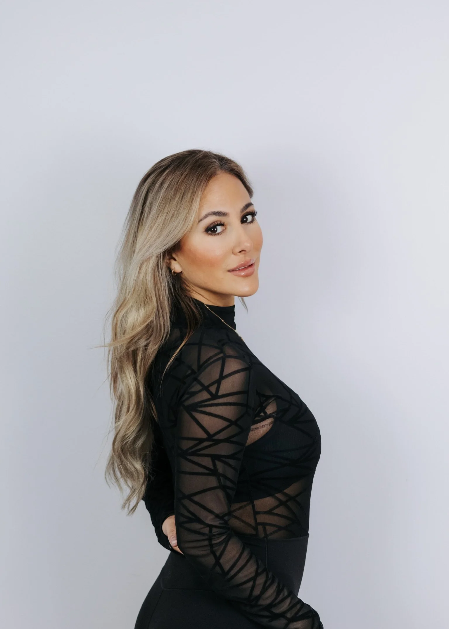 A woman with long, wavy blonde hair and makeup, wearing a black, sheer, patterned top and black pants, standing against a plain white background, looking at the camera.