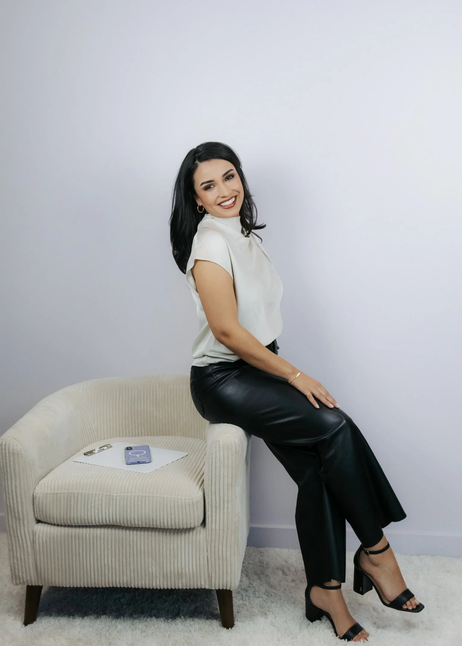 A woman with black hair, wearing a white top, black leather pants, and black high heels, sitting on a beige armchair with a smartphone and glasses placed on the chair next to her, smiling at the camera against a plain light-colored wall.