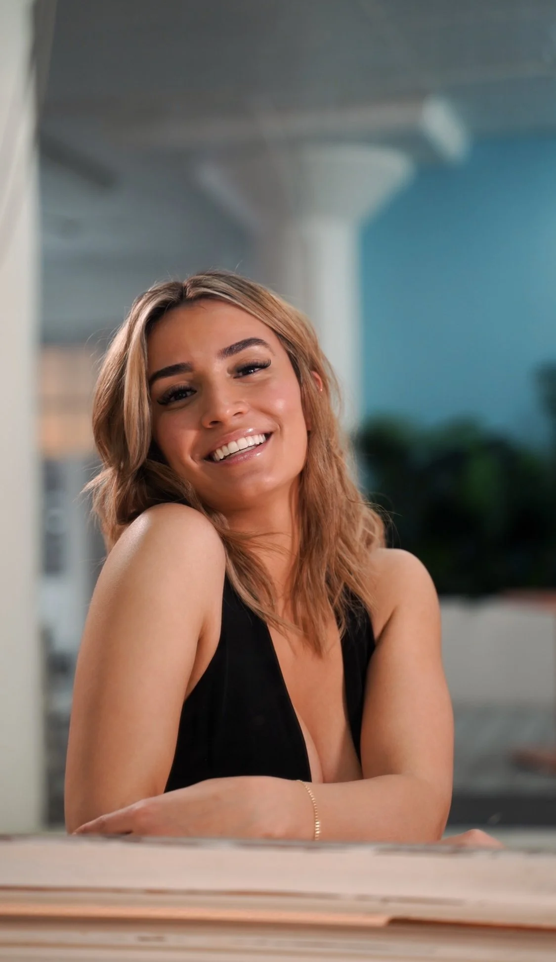 A young woman with shoulder-length light brown hair, smiling and wearing a sleeveless black top, sitting at a table indoors with a blurred background of an interior space and some outdoor elements.