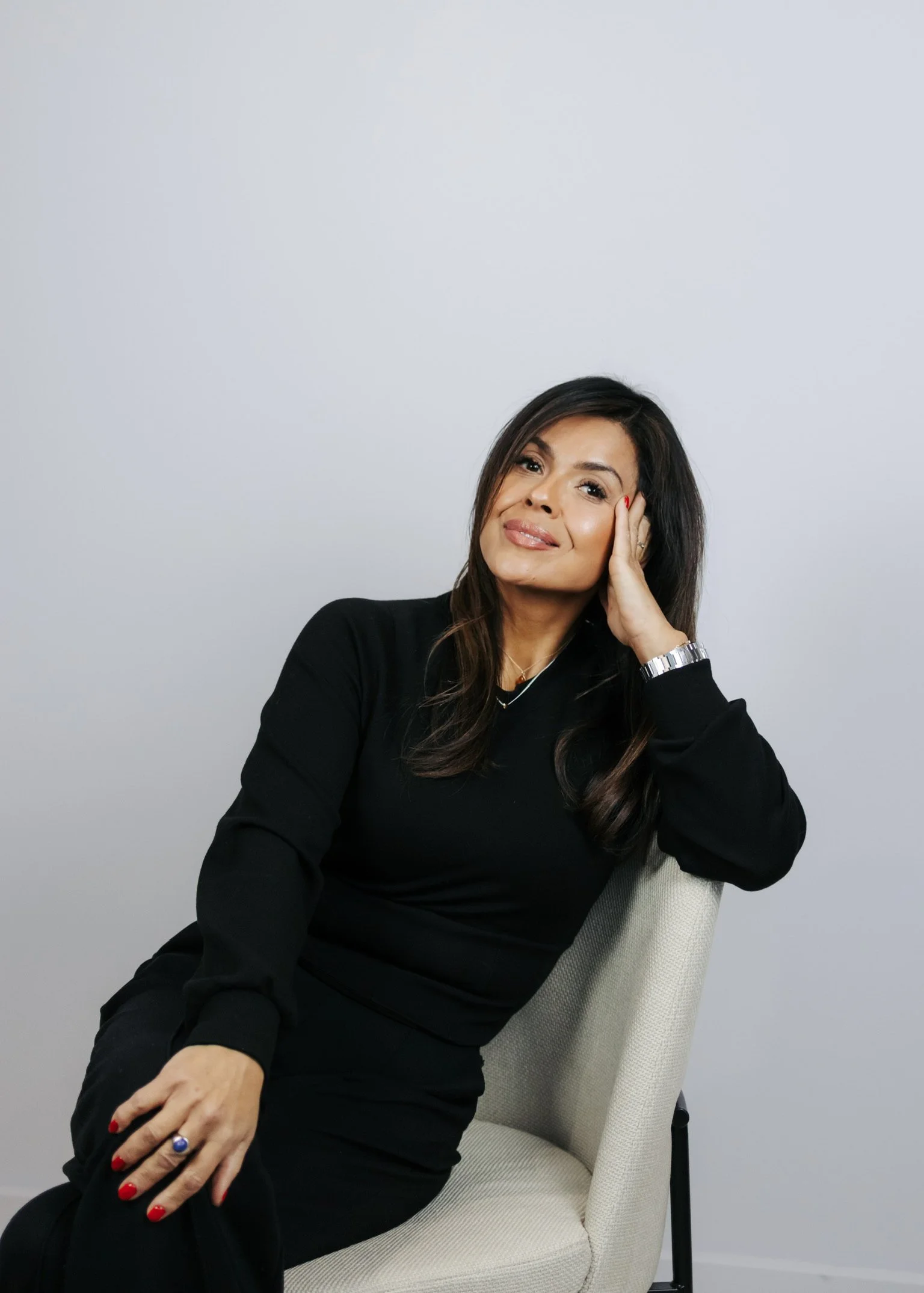 A woman with long dark hair, wearing a black top and black pants, sitting on a light-colored chair, smiling with her head resting on her hand against a plain white background.