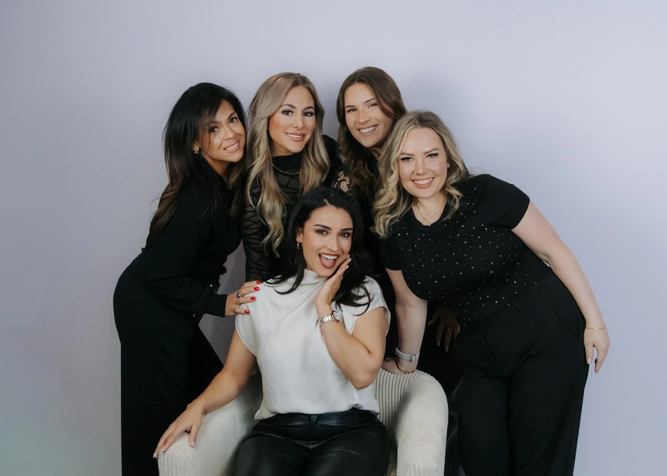 Six women posing together, smiling, with one sitting in a chair in front and five standing behind her, against a light gray background.
