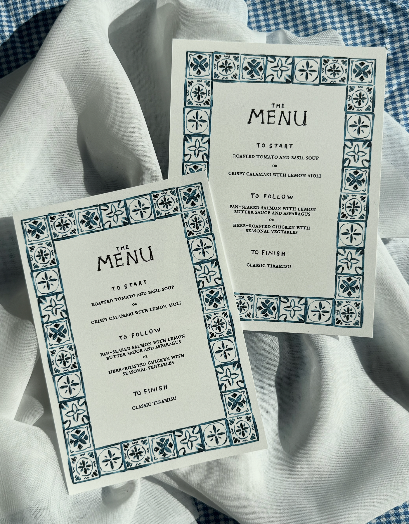 Two menus with blue floral border designs, listing meal options including soup, calamari, salmon, chicken, and tiramisu, placed on white linens.