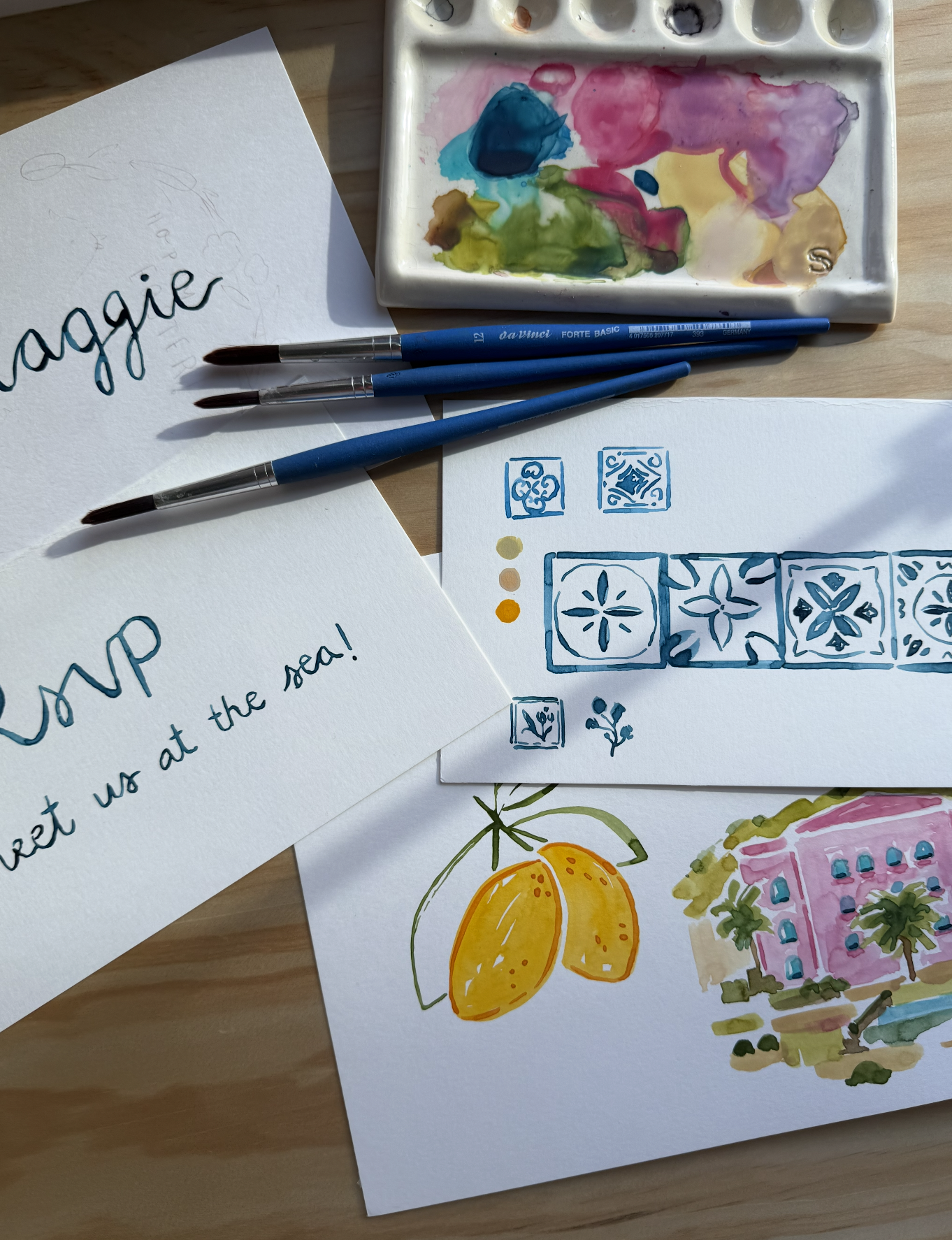 Watercolor painting supplies and handmade notes on a wooden table. The supplies include a watercolor palette with mixed colors, three blue paintbrushes, and illustrations of a lemon, decorative tile patterns, and a pink building with palm trees. Hand