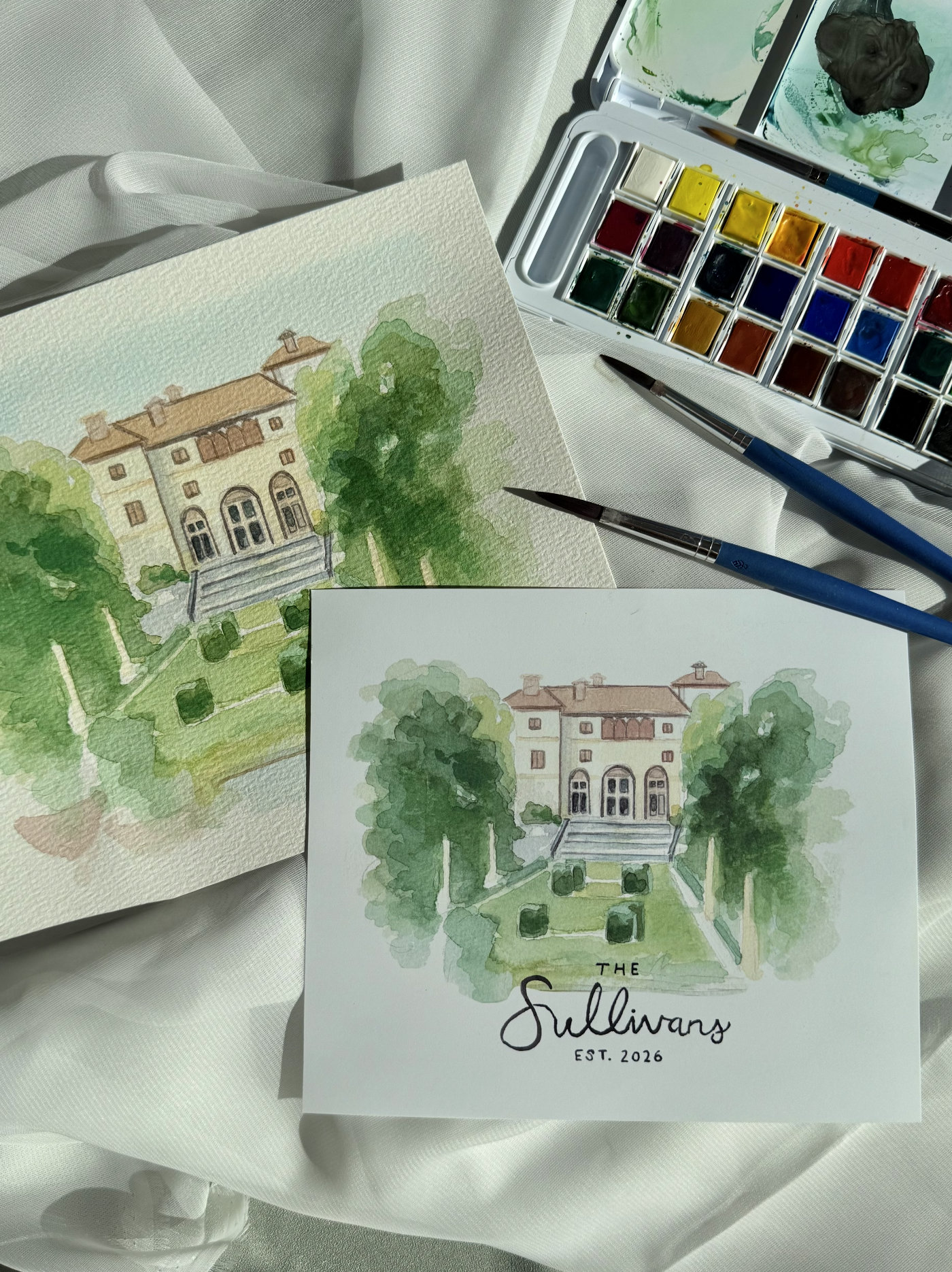 Watercolor painting of a large house with trees and a landscaped garden, with a branded sign reading 'The Sullivans Est. 2026' placed in front of the painting. Art supplies including a watercolor palette, brushes, and a palette knife are visible on a