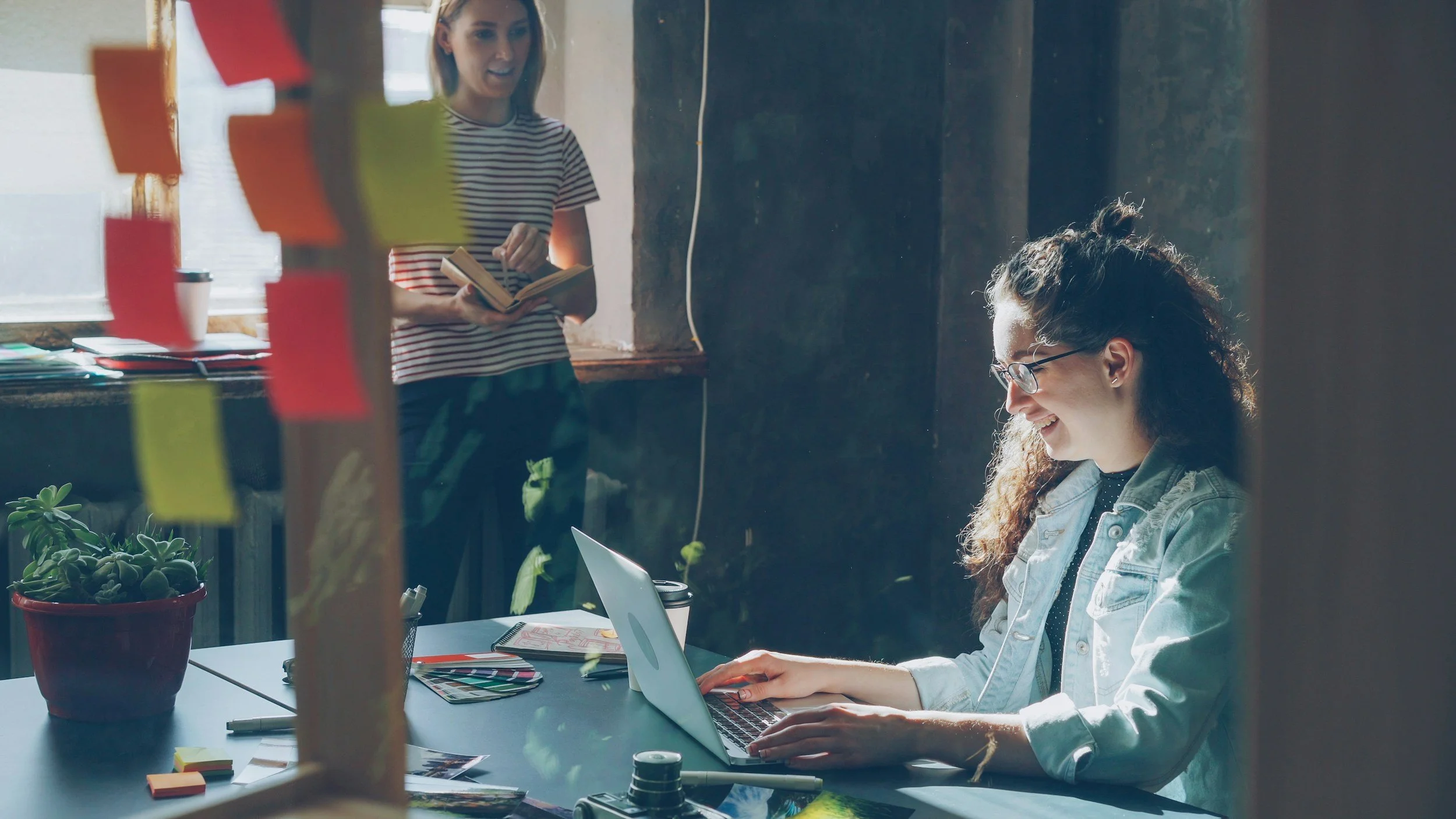 Two women working in a modern office with a large window, sticky notes, plants, a laptop, and office supplies on the desk.