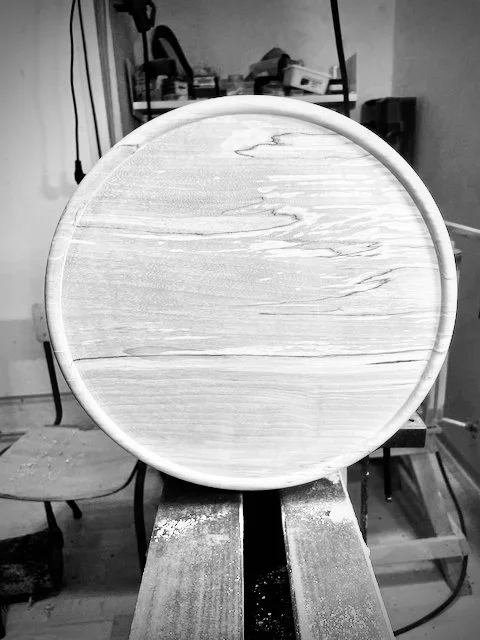 A round wooden tabletop on a wooden workbench in a woodworking shop.