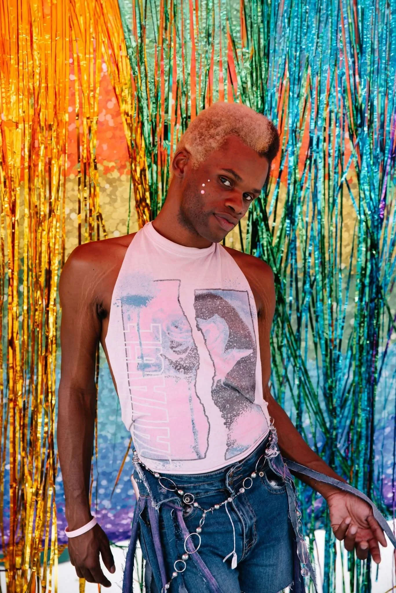 A person with bleached blonde hair and dark skin standing in front of a colorful background made of shiny, metallic streamers in orange, green, blue, and purple colors. The person wears a sleeveless graphic tank top, ripped jeans with chains, and has face paint with small white dots on their cheek.