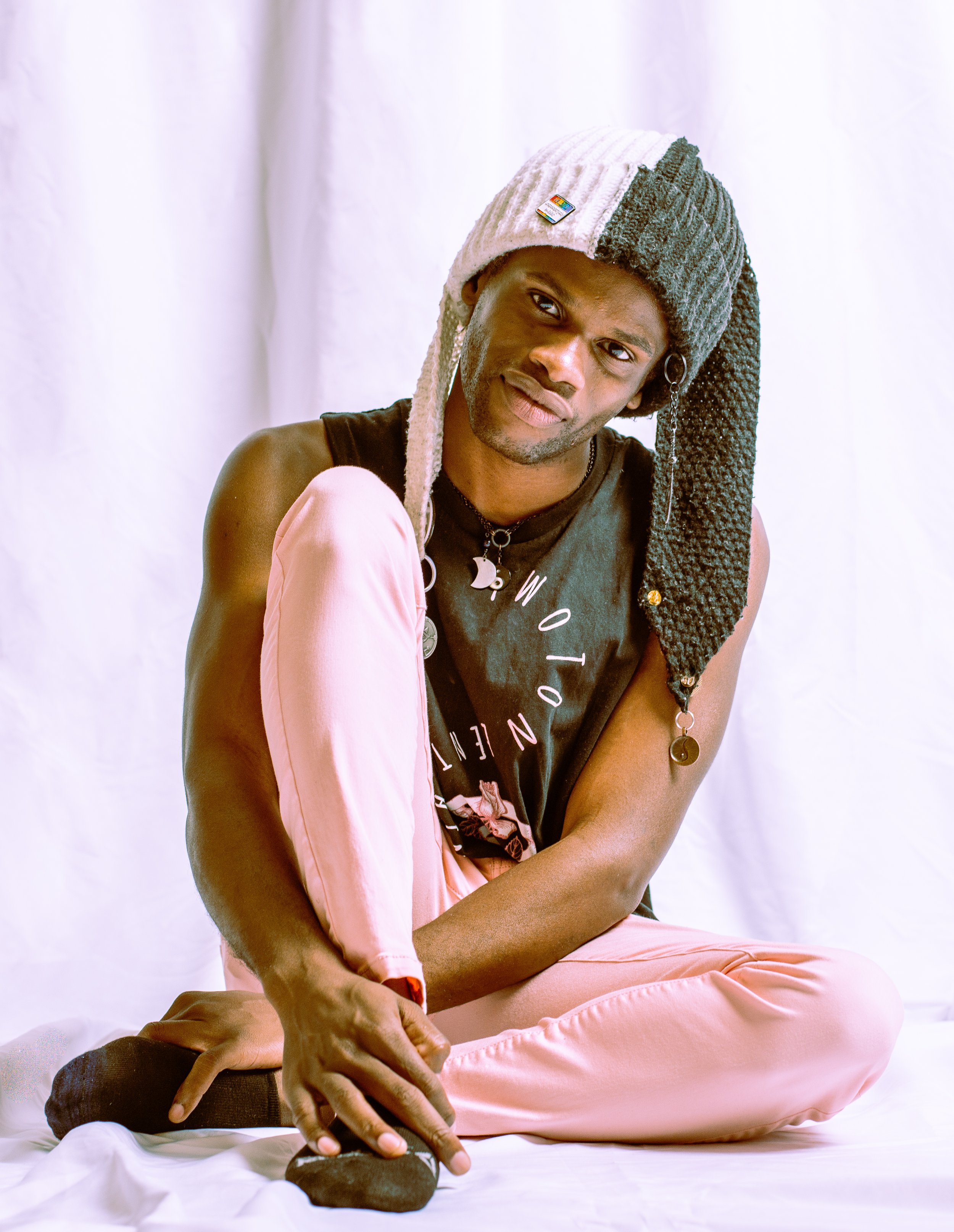 A person sitting cross-legged on the floor, wearing a gray and pink knit hat, a sleeveless black shirt, pink pants, and black socks, with a white background.