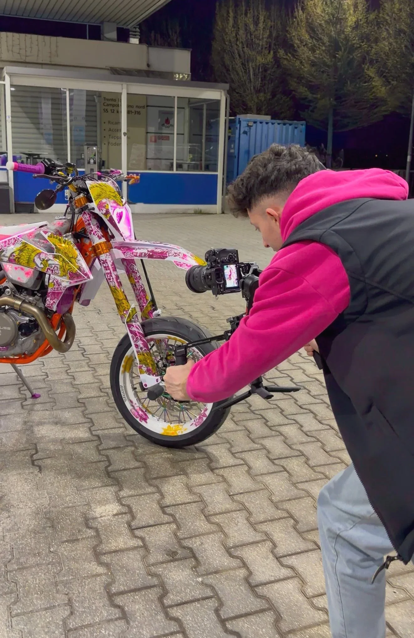 Persona che fotografa una moto colorata con fiori. È sera, in un'area esterna, probabilmente un parcheggio, con un edificio di servizio e alberi sullo sfondo.