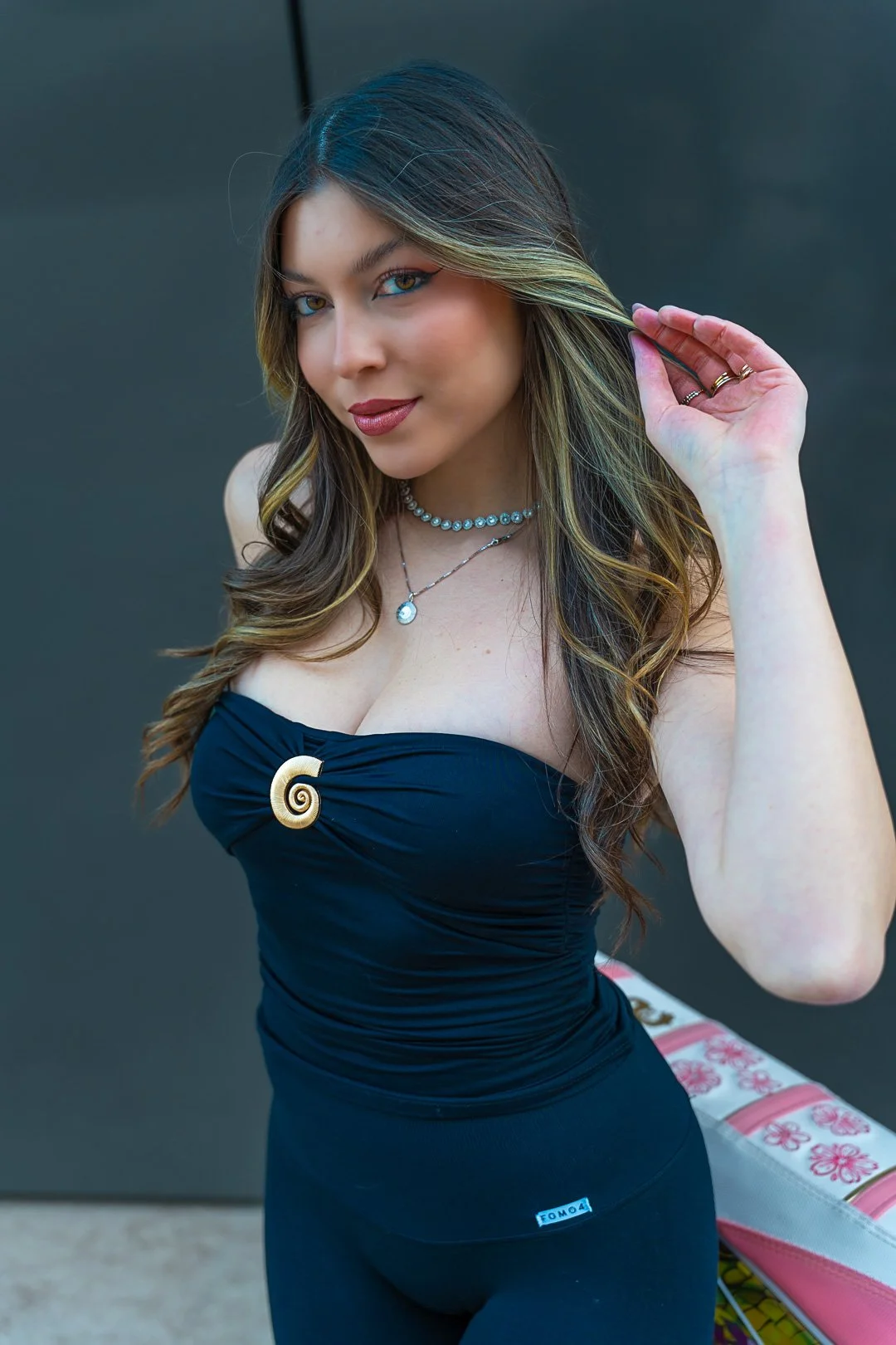 Donna con capelli castani, con capelli biondi con tinte di castano, che indossa una collana di perle, una collana con ciondolo, e una maglietta nera con una spirale dorata come decorazione.