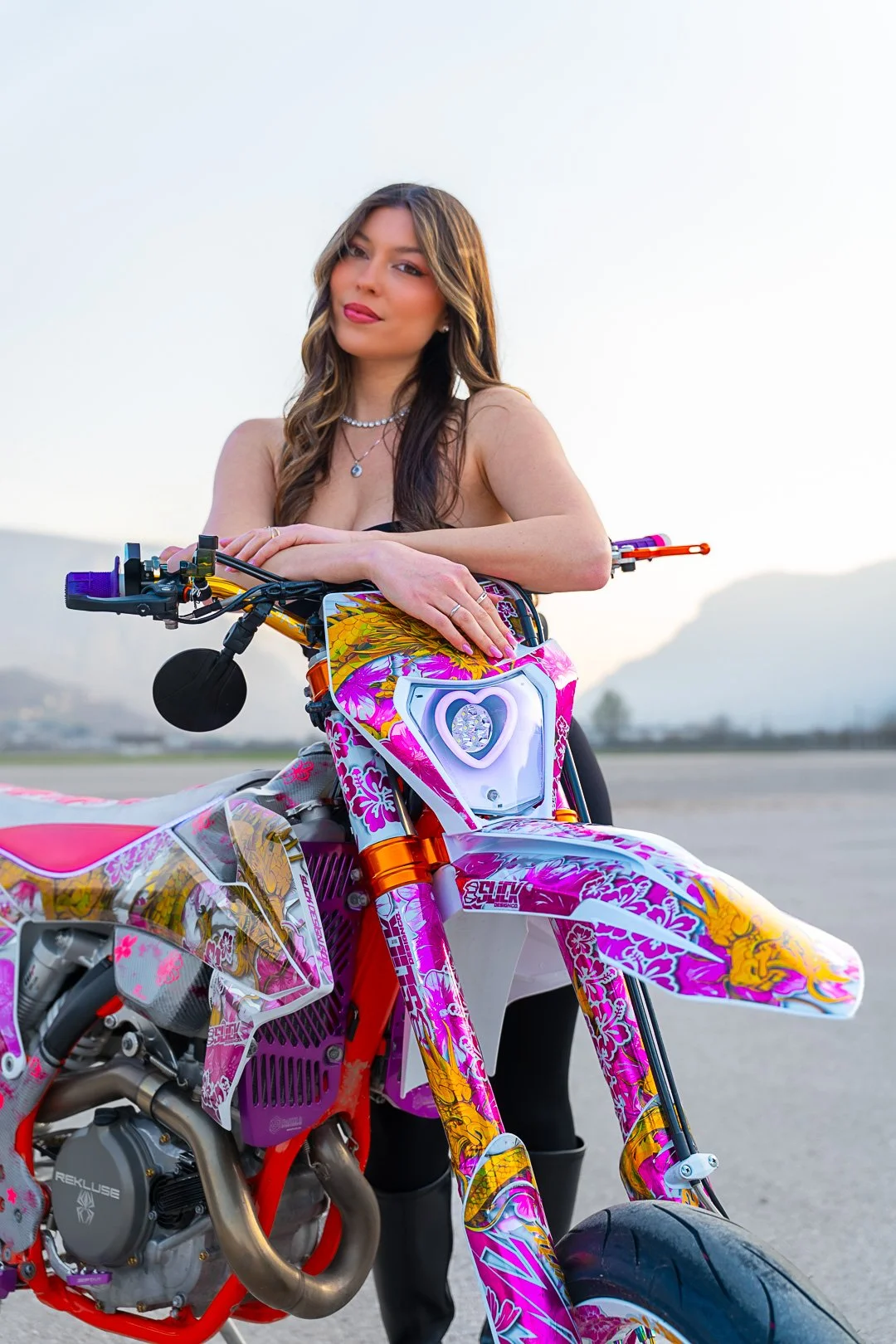 Una donna con capelli castani e occhi chiari, indossa gioielli e si appoggia su una moto da cross decorata con fantasie rosa, viola e gialle, in un ambiente aperto con montagne sullo sfondo.