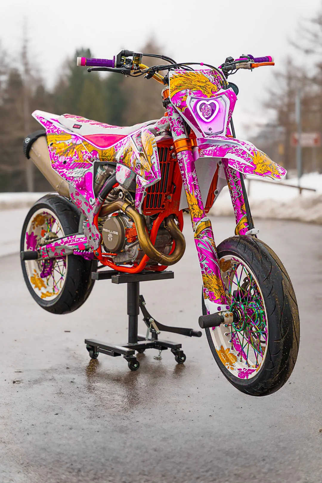 Motocicletta da trial decorata con colori vivaci, principalmente rosa, giallo e bianco, in posa su un supporto su una strada.