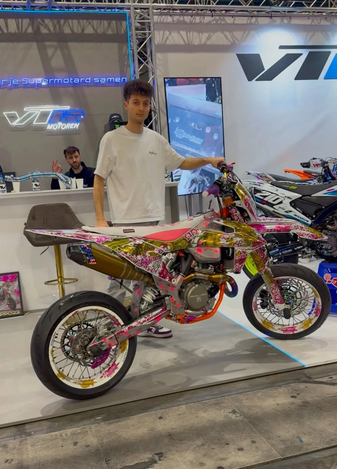 Persona che mostra una moto decorata con motivi floreali rosa e dorati in un exhibit con altre moto in background.