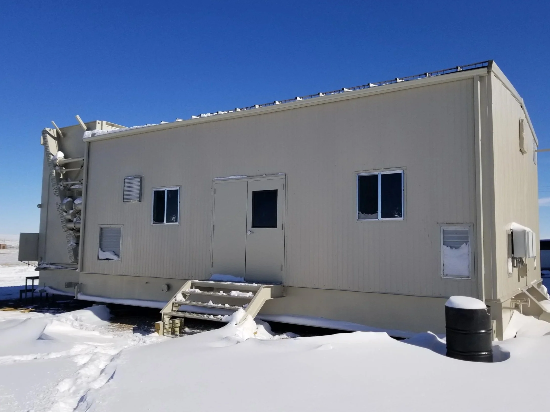 A beige modular building in a snowy landscape with clear blue sky, featuring small stairs leading to a door and several windows, some covered with snow.