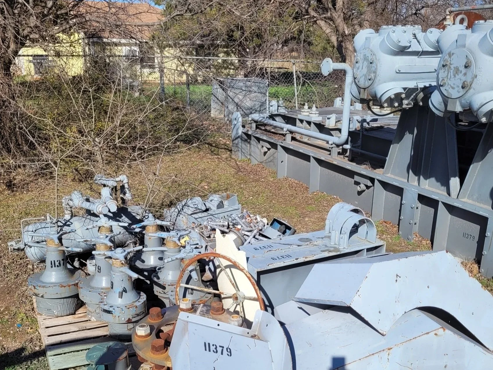 Collection of old, rusted mechanical and electrical equipment outdoors, including large gray transformers, gauges, pipes, and small devices on a wooden pallet, with trees and a chain-link fence in the background.