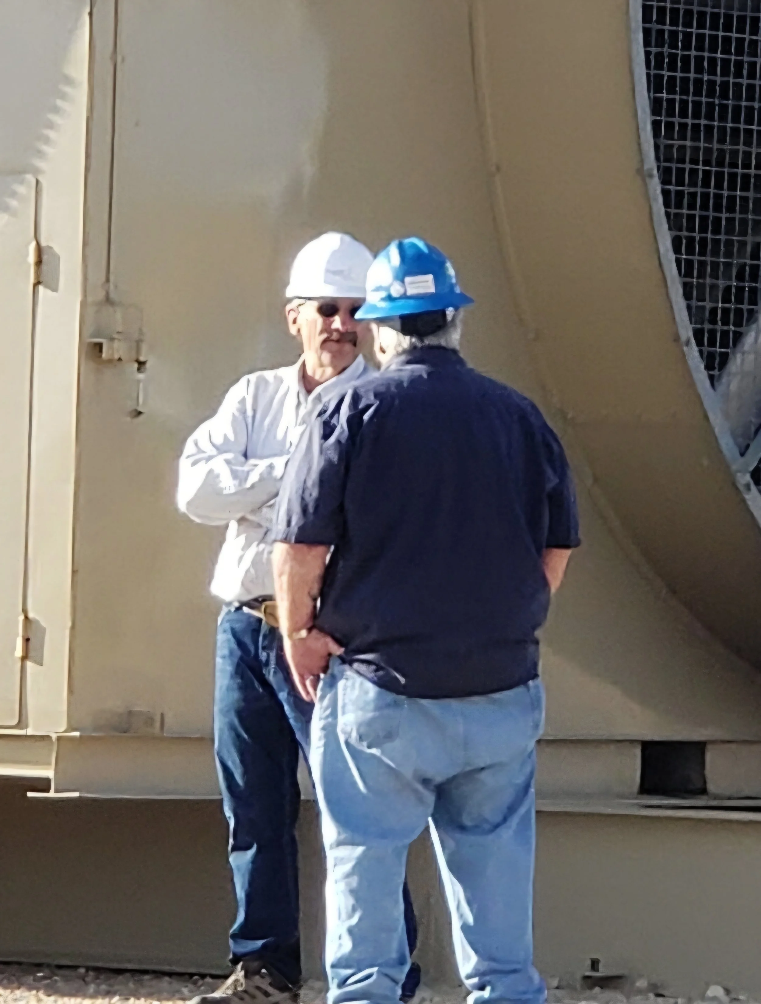 Two men with hard hats, one in white and one in blue, talking outdoors near industrial equipment.