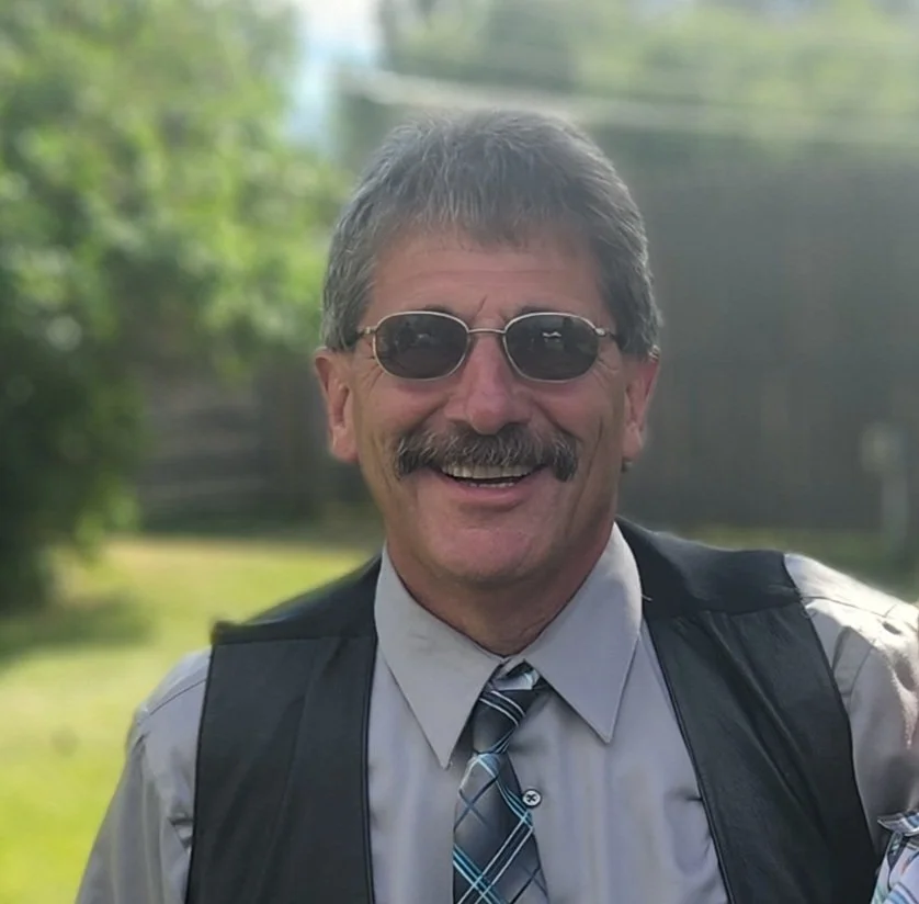 A middle-aged man with a mustache and gray hair wearing sunglasses, a light dress shirt with a striped tie, and a black vest, smiling outdoors with greenery in the background.