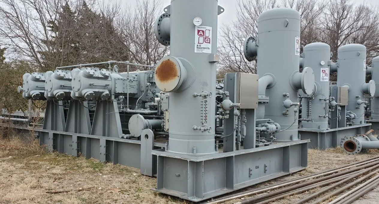 Industrial equipment with large gray cylindrical tanks, pipes, valves, and control boxes outdoors on a metal platform, with leafless trees in the background.