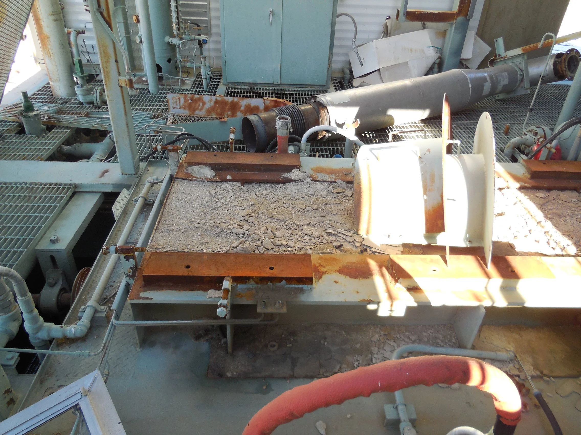 Close-up of an industrial outdoor machinery area with rusted metal parts, pipes, and equipment, including a large gray pipe and a rusted metallic surface with dirt and debris.