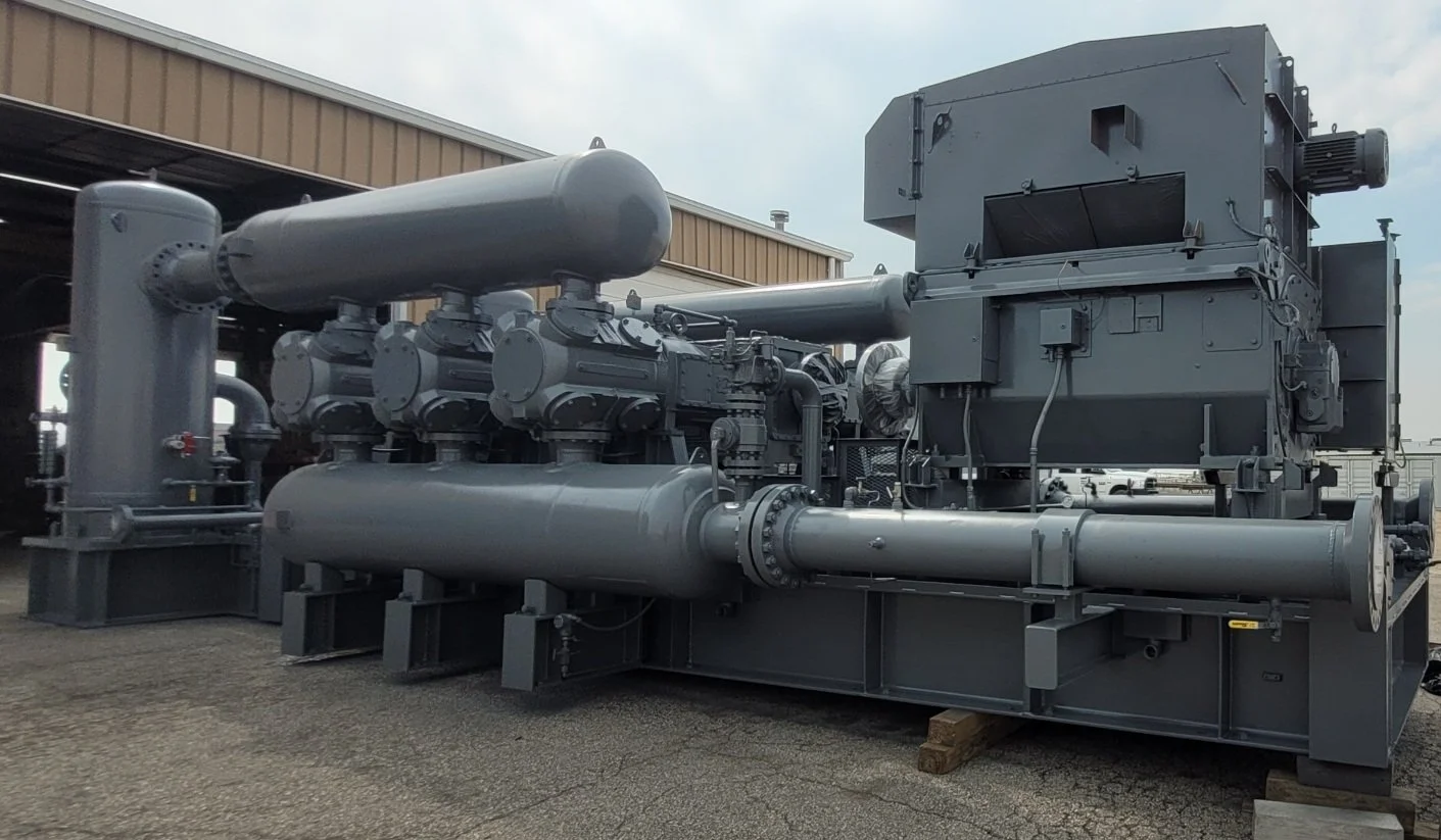 Large industrial compressor or pump system painted in gray, installed outdoors on concrete ground, with a building and cloudy sky in the background.
