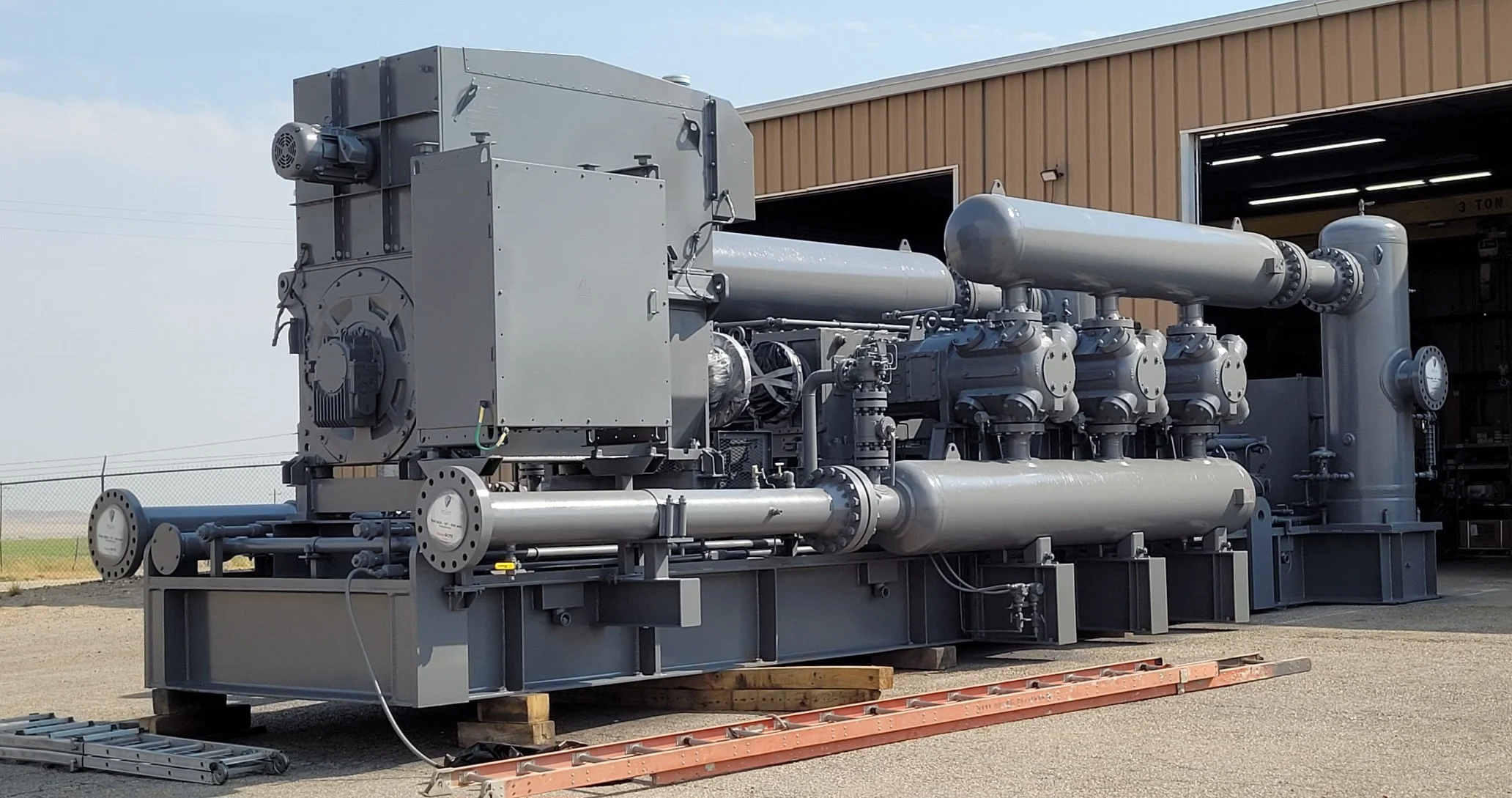Large industrial machine or equipment outdoors, with multiple pipes, valves, and a control box, near a building with a garage door, placed on wooden pallets and surrounded by clear sky and a fence.